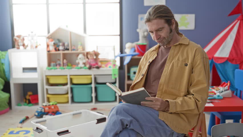 Man yawning with arms raised while holding a tablet amid scattered toys in a building classroom; parental exhaustion.