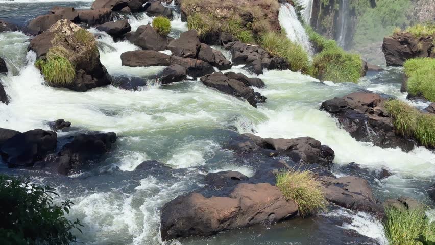 Close-up of churning white water rapids at the top of a massive waterfall system with lush green plants growing on volcanic rocks under the bright sun