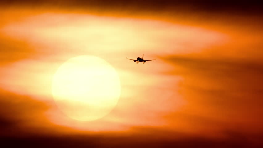 Passenger jet silhouette flying across a massive glowing sunset sun. Warm orange sky with hazy clouds during dusk, captured with extreme telephoto zoom lens.