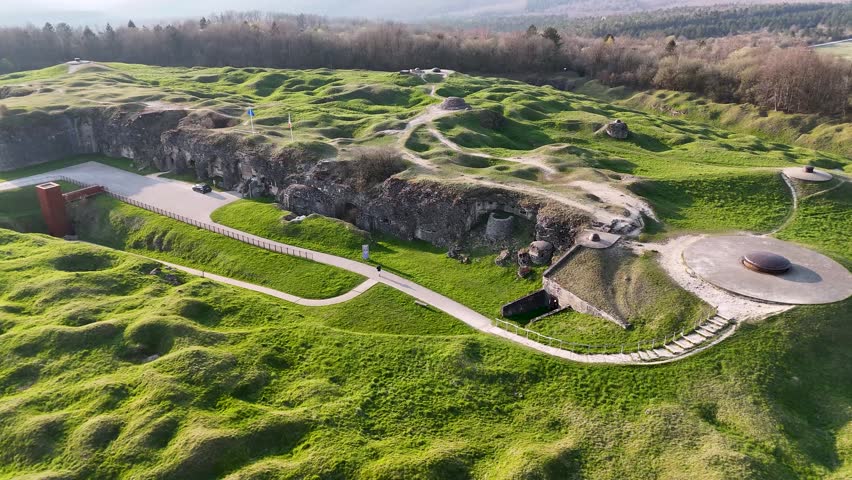Drone perspective of sunlit Fort Douaumont earthworks, paths, and woodland on the Verdun battlefield in Grand Est, France.