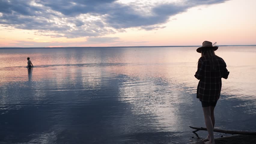 Silhouette of a young female photographer in a hat taking pictures of a beautiful sunset over the calm sea while a man swims in the distance, creating a serene and picturesque landscape scene