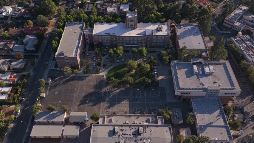 Top down orbit shot showcasing historic brick school building with clock tower, playground, basketball court in residential neighborhood of Los Angeles, California