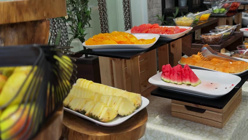 Bright daytime view of a fresh sliced fruit buffet on white platters and tiered wooden stands. The colorful healthy breakfast display features juicy pineapple, watermelon, and oranges at a hotel.