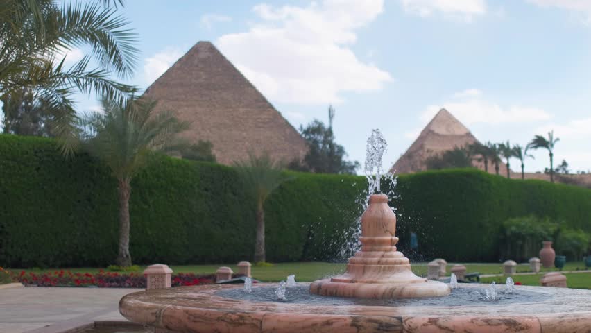 Slow motion 4K travel video featuring a fountain in the foreground with the Pyramids of Giza in the background, surrounded by palm trees and lush gardens in Egypt.