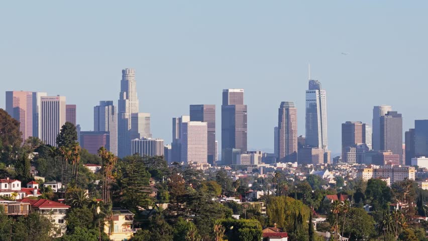 Scenic flight revealing downtown Los Angeles skyscrapers emerging behind Hollywood Hills residential buildings with lush green trees