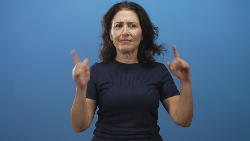 Woman in navy shirt pressing fingers into ears and grimacing in studio blue backdrop; sensory overload discomfort.