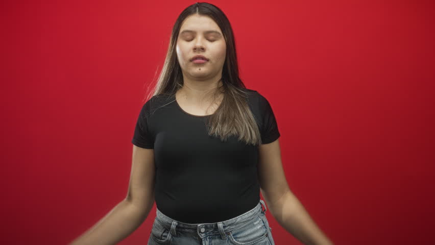Young woman smiles and points both index fingers downward at camera, wearing black tee and jeans in a red studio; playful invitation.