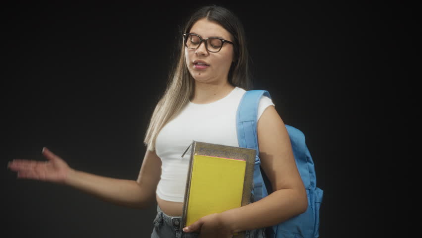 Young woman wearing white crop top and denim jeans holding books and blue backpack, hand covering eyes gesture in studio; playful.