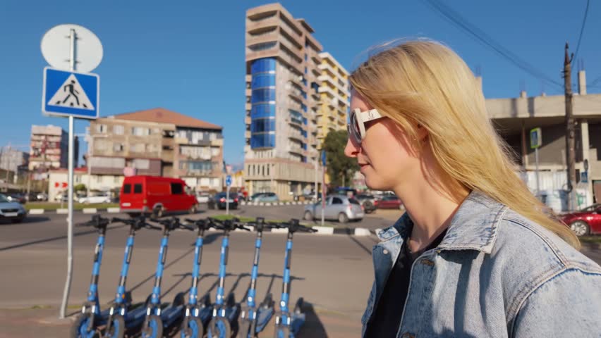 Young blonde woman wearing a denim jacket and stylish sunglasses walking along a busy city street on a sunny day, with her hair blowing in the wind as traffic passes in the background