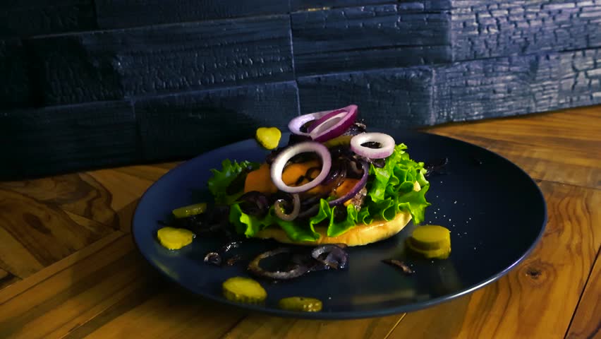 Professional cook wearing black gloves putting the top bun on a gourmet hamburger served on a dark plate, carefully completing the preparation of the delicious fast food meal in a restaurant