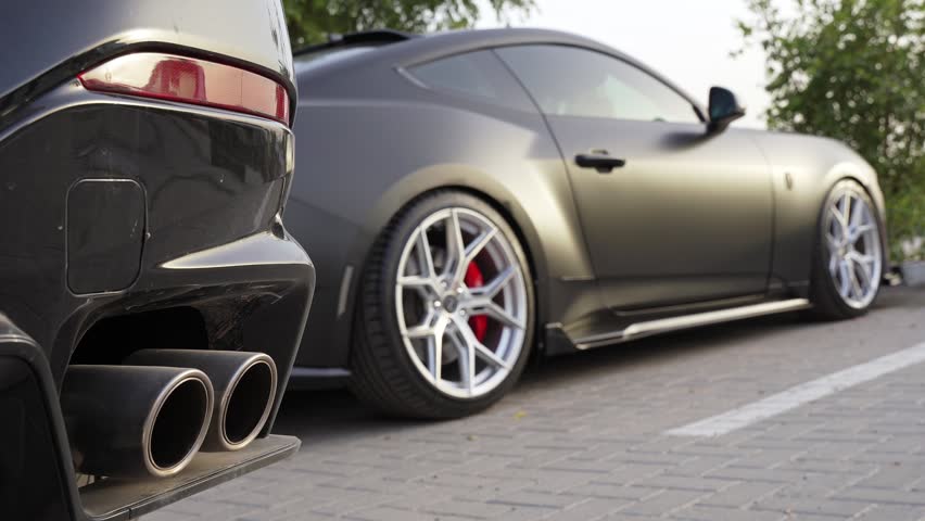 A low-angle close-up shot focusing on the dual exhaust pipes of a black vehicle, with a custom matte black sports car parked on a paved lot blurred in the background.