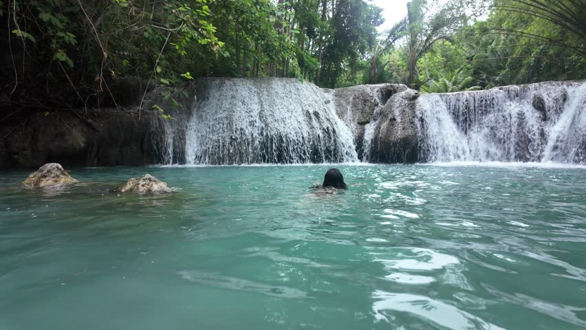 Woman swimming in turquoise waterfall lagoon surrounded by tropical jungle