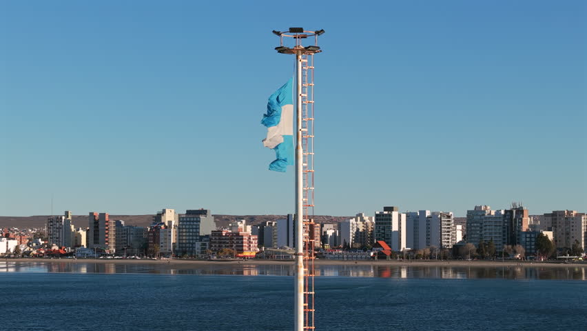 Argentine national flag waving above the waterfront of Puerto Madryn, Chubut, Argentina, with Patagonian coastal skyline and Atlantic waters in background, slow motion aerial shot.