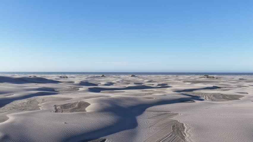 Wide view of vast rippled coastal sand dunes extending to calm ocean horizon under clear blue sky in New Zealand