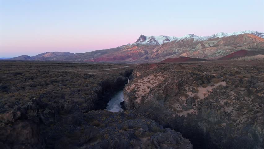 Stunning drone footage of a river flowing through a dark volcanic canyon at sunrise in La Payunia Reserve, Mendoza, Argentina. Beautiful pink sky.