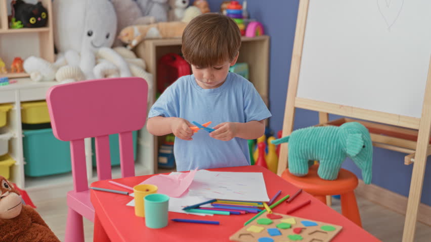 Boy cutting paper with scissors at a small red table, holding sheet with hands, surrounded by crayons and stuffed toys in a building playroom; concentration creativity.