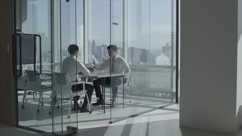 Two Asian businessmen in white dress shirts and ties shaking hands and smiling inside a glass-walled meeting room in a modern high-rise office building, viewed from the corridor outside, with a bright city skyline visible through floor-to-ceiling windows. Korean text "회의실4" on office glass wall. Translation: "Meeting Room 4".