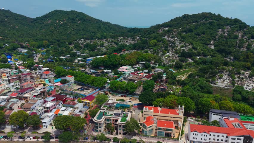 Aerial parallax above riverfront promenade in Coloane Village Macau revealing ancient houses, colorful rooftops, hillside cemetery and forested hill with sea behind
