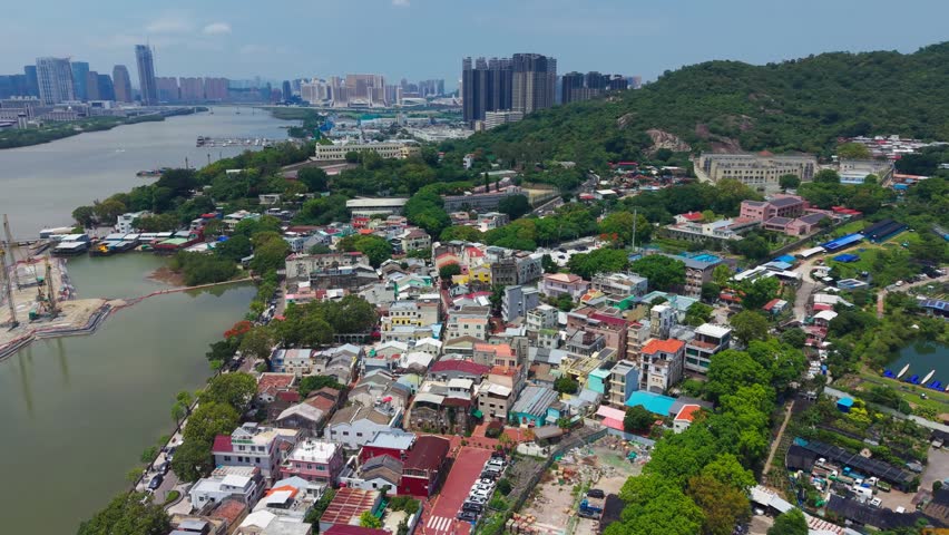 Aerial flyover of Coloane Village in Macau showing traditional low-rise houses by river channel with modern skyscraper skyline and green hillside behind