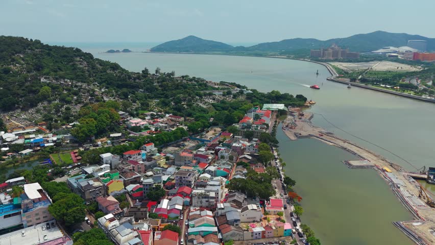 Aerial parallax view over Coloane Village in Macau showing colorful rooftops, forested hillside, waterfront promenade and calm sea with distant islands