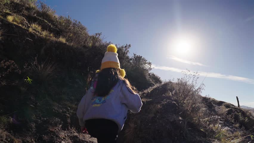 A young child wearing a colorful beanie hikes up a steep rocky mountain trail under bright sunshine.