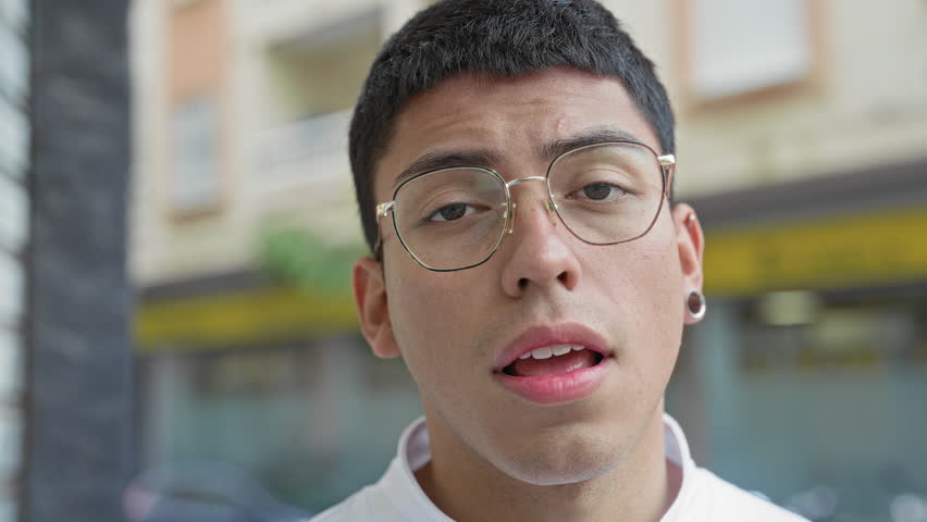 Man with glasses gazes at camera, lips slightly parted and one eye half closed, earring visible on a street; pensive.