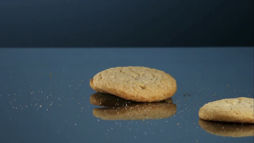 Delicious round sugar cookies falling and bouncing on a dark blue reflective surface in slow motion with crumbs scattering for bakery and dessert advertising