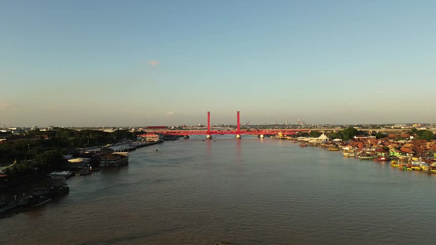 Aerial drone shot flying over the Musi River towards the iconic Ampera Bridge in Palembang, South Sumatra, Indonesia. Traditional boats and urban cityscape at sunset
