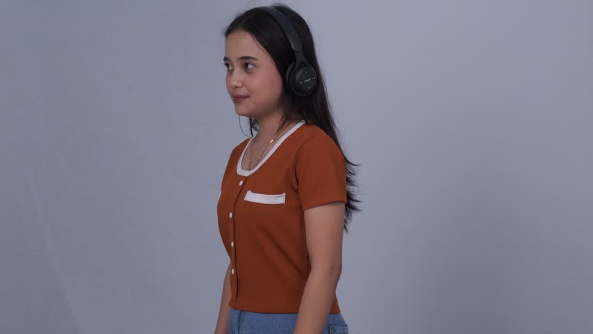 A young Asian woman wearing an orange t-shirt and jeans is smiling beautifully and wearing a black headset while enjoying music, seen from the side.