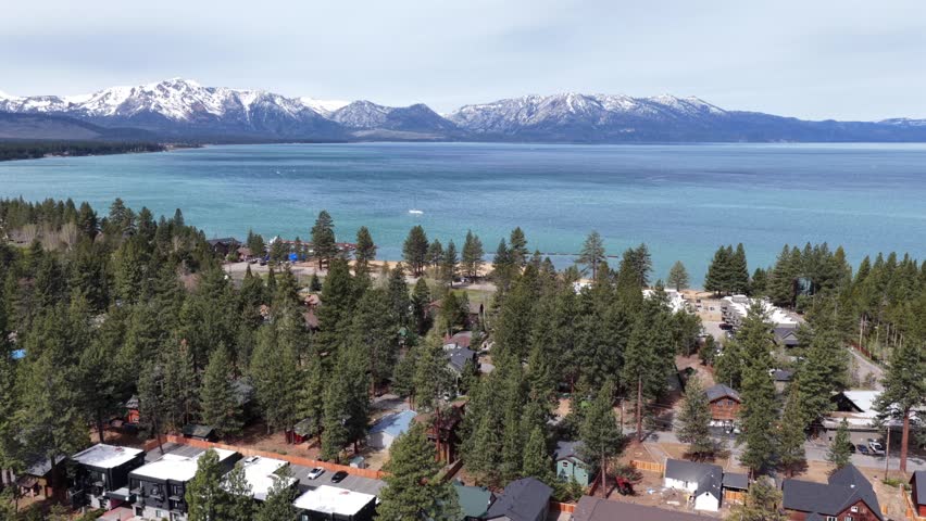 Lake Tahoe shoreline with alpine cabins and pine forest in foreground, turquoise lake and snow-capped Sierra Nevada peaks filling horizon under overcast skies. California, USA. Aerial flyover shot.