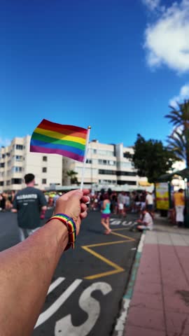 Point of view of a person holding a small waving LGBTQ+ rainbow flag during a sunny gay pride parade on a city street, showing support for the community and celebrating diversity and equality