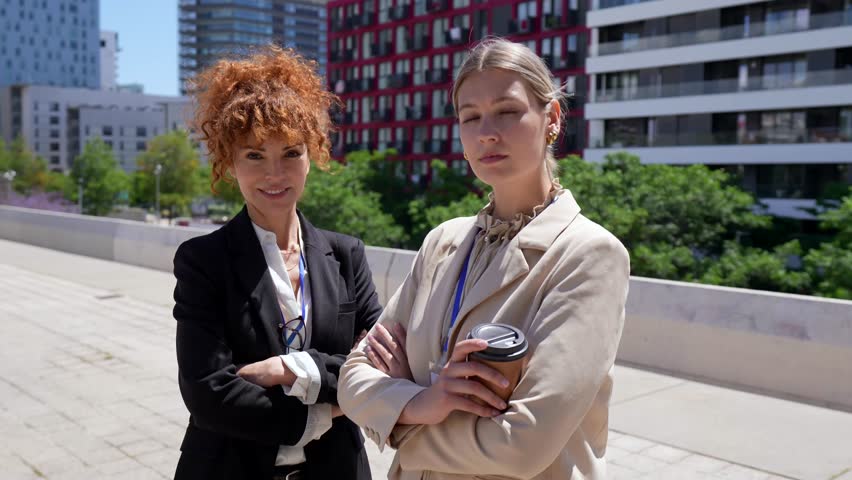 Two determined businesswomen, a mature redhead and a young blonde colleague, standing outdoors with arms crossed, projecting confidence and success against a modern architectural background