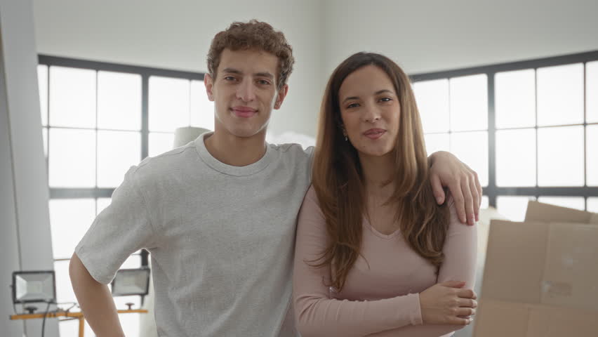 Man with arm around woman gesturing with open hands beside moving boxes in building; moving day joy and togetherness.