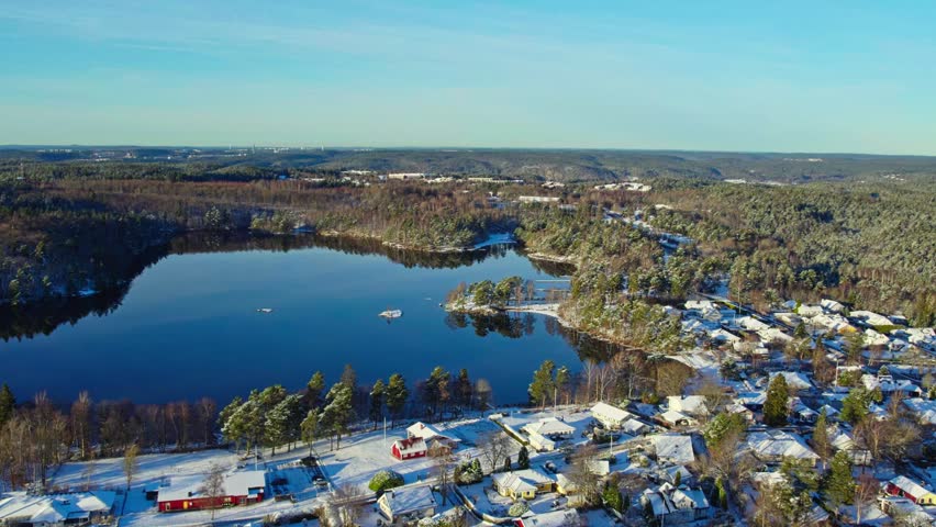 Aerial view of snow-covered houses and forest surrounding calm lake in winter daylight