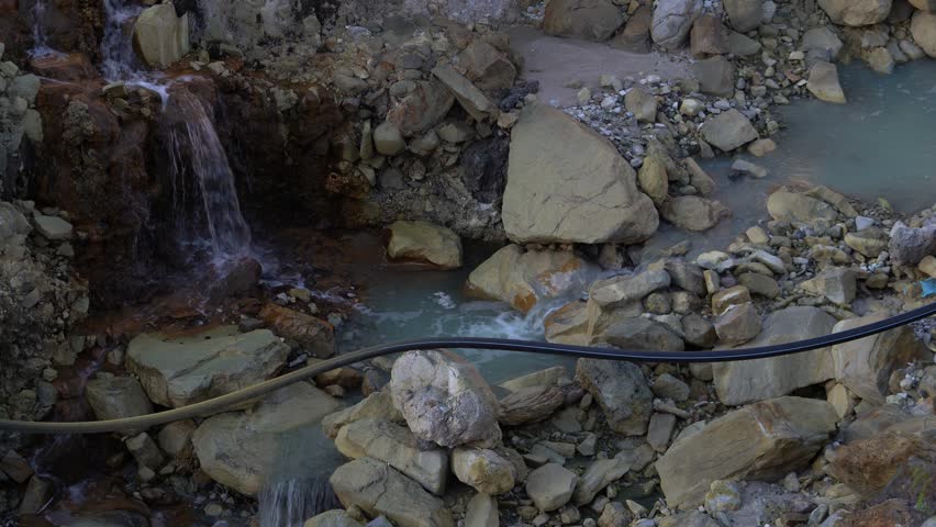 A small waterfall flowing over rocky terrain in a natural geothermal area with mineral deposits and a black pipe running across the scene