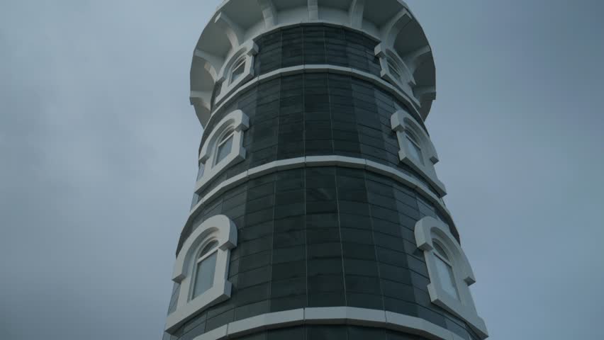 A handheld camera circles the exterior of a tiled lighthouse at Lake Baikal from a low angle. The close-up shot highlights the white arched windows and dark facade against an overcast sky.