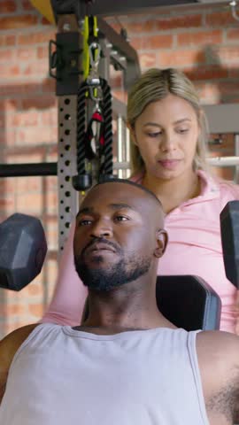 Vertical video: Lifting man bracing hex dumbbells on incline bench in gym, tank top, spotter cuing. Weights, strength, workout, partner, coaching, focus, vertical video