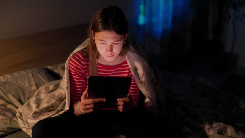 Young girl experiencing insomnia sitting in bed at night browsing on a digital tablet. Colored blue and red ambient neon light illuminates her face.
