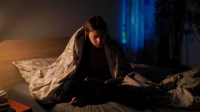 Tired young girl yawning and closing her book while sitting in bed struggling with late night insomnia and fatigue. Sleepy child lifestyle in bedroom.