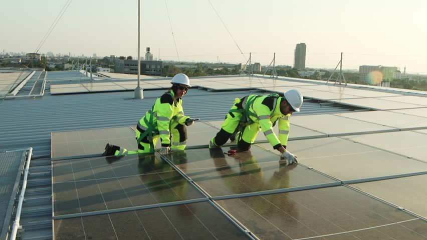 Two service engineers in safety clothing with helmet check solar system at rooftop solar farm with city view. Engineers installing solar panels. Man and woman workers inspecting solar panels on roof.