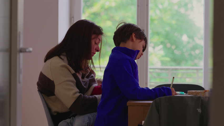 Woman leaning close to child while he writes in notebook near window conveying concentration, encouragement and nurturing everyday family education routine