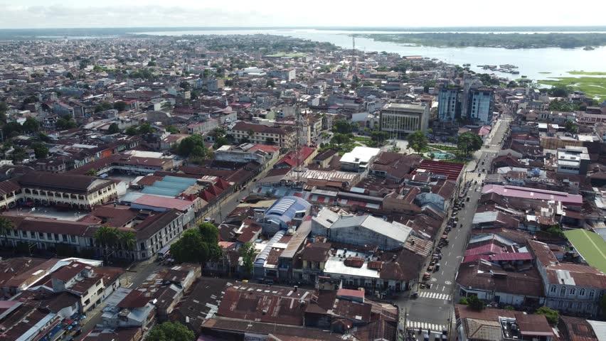 An expansive aerial shot capturing the bustling cityscape of Iquitos, Peru, featuring a blend of traditional red-roofed buildings and contemporary structures against vivid natural surroundings, including the Amazon River in the distance.