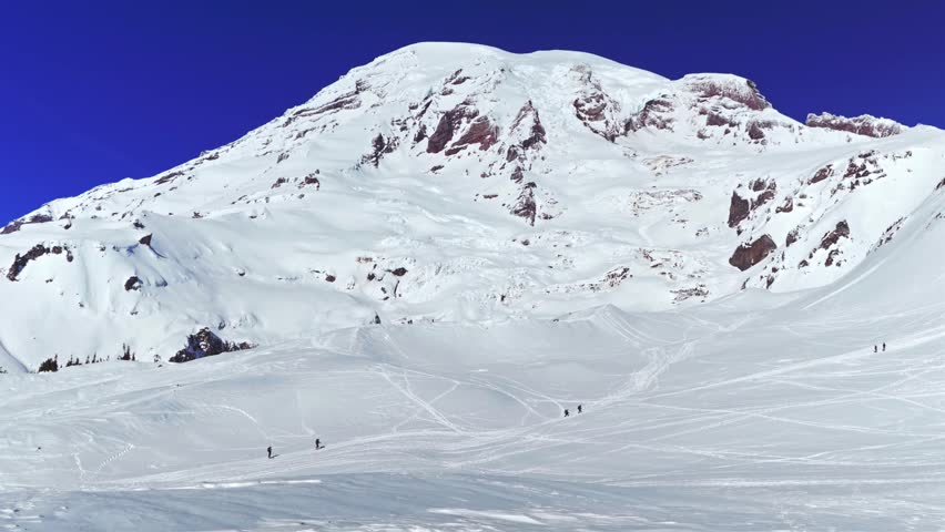 Scenic view of the snow-covered Mount Rainier summit and surrounding landscape in Mount Rainier National Park, Washington State, USA. The footage captures individuals snowshoeing or backcountry skiing