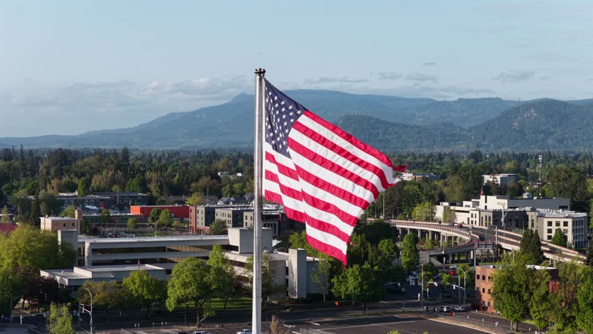 Large American flag waving dramatically on tall flagpole above Eugene downtown, low-rise commercial buildings and tree-lined streets with forested hills beyond. Oregon, USA. Aerial orbit shot.