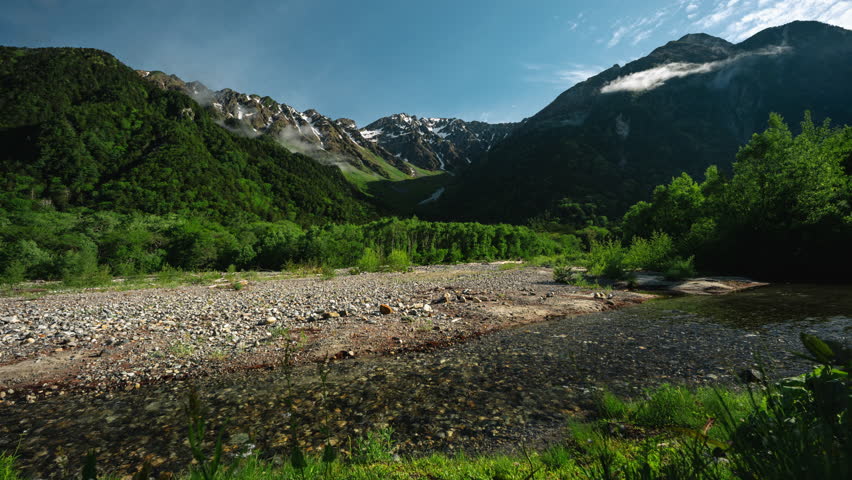 Japan Alps Kamikochi in Chubu Sangaku National Park Mountain valley river landscape with green forest, rocky shore, snowy peak, blue sky, summer sunlight, tranquil nature, wide view, wilderness scene