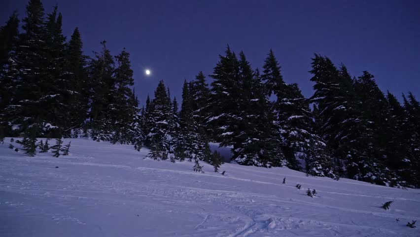 Evening or night footage of a snowy slope under a dark blue sky with a bright moon, along the Skyline Trail towards Panorama Point in Mount Rainier National Park. Tracks in the snow suggest winter rec