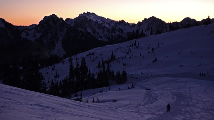Footage captures a lone snowshoer hiking up the snow-covered Skyline Trail in Mount Rainier National Park during a beautiful winter sunrise or sunset. The trail leads towards Panorama Point with the r
