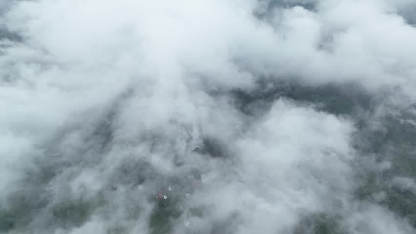 Majestic aerial view flying forward over a sea of white clouds, revealing a lush green tropical forest and a small village nestled on the mountainside below in a foggy, atmospheric scene