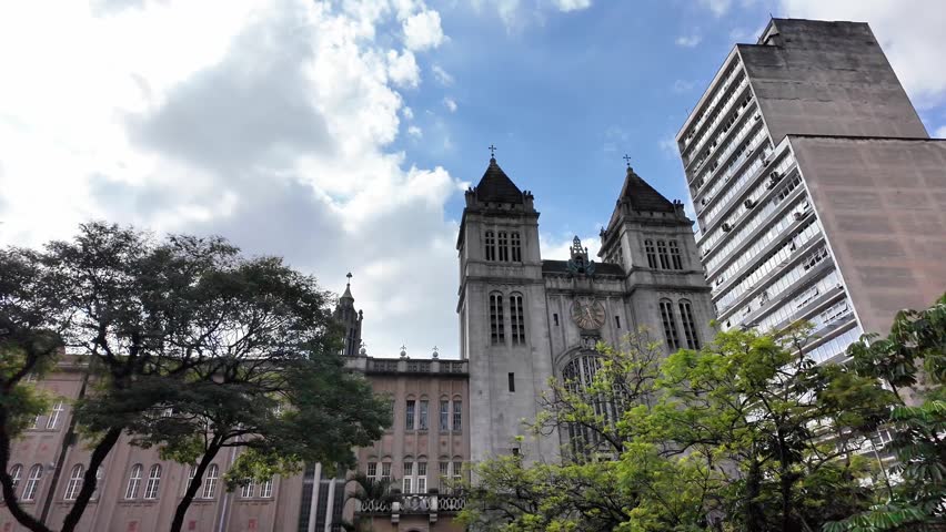 Facade of Mosteiro de Sao Bento and Modern Skyscraper in Sao Paulo, Brazil; High-quality footage featuring a low-angle shot of the historic Mosteiro de Sao Bento in the heart of downtown Sao Paulo, Brazil. The impressive stone facade, complete with its large ornate window and twin towers, stands in striking contrast to the modern skyscraper adjacent to it. A subtle sun flare adds a cinematic touch to this iconic religious and architectural landmark. Ideal for travel documentaries, historic urban features, and content focusing on religious architecture.