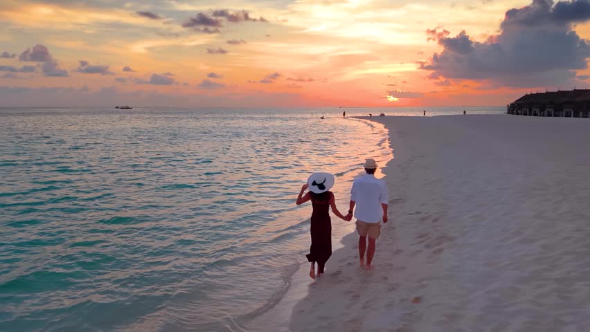 Slow motion aerial view of a romantic couple holding hands and walking down a beautiful beach in the Maldives islands during sunset time